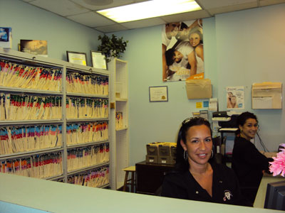 The image shows an indoor office environment with a woman seated at a desk, a reception area with a counter and a bulletin board displaying various items, and shelves containing files.