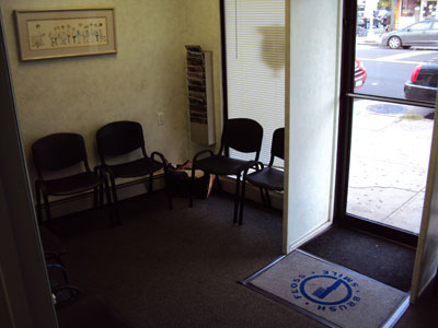 The image shows an interior view of a waiting room with multiple chairs arranged around a table, leading towards a door with a glass panel and a floor mat.