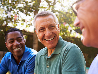 The image depicts three older men sitting outdoors, smiling and engaging with each other.