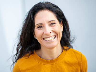 The image shows a smiling woman with long hair, wearing a yellow top, against a blurred background.