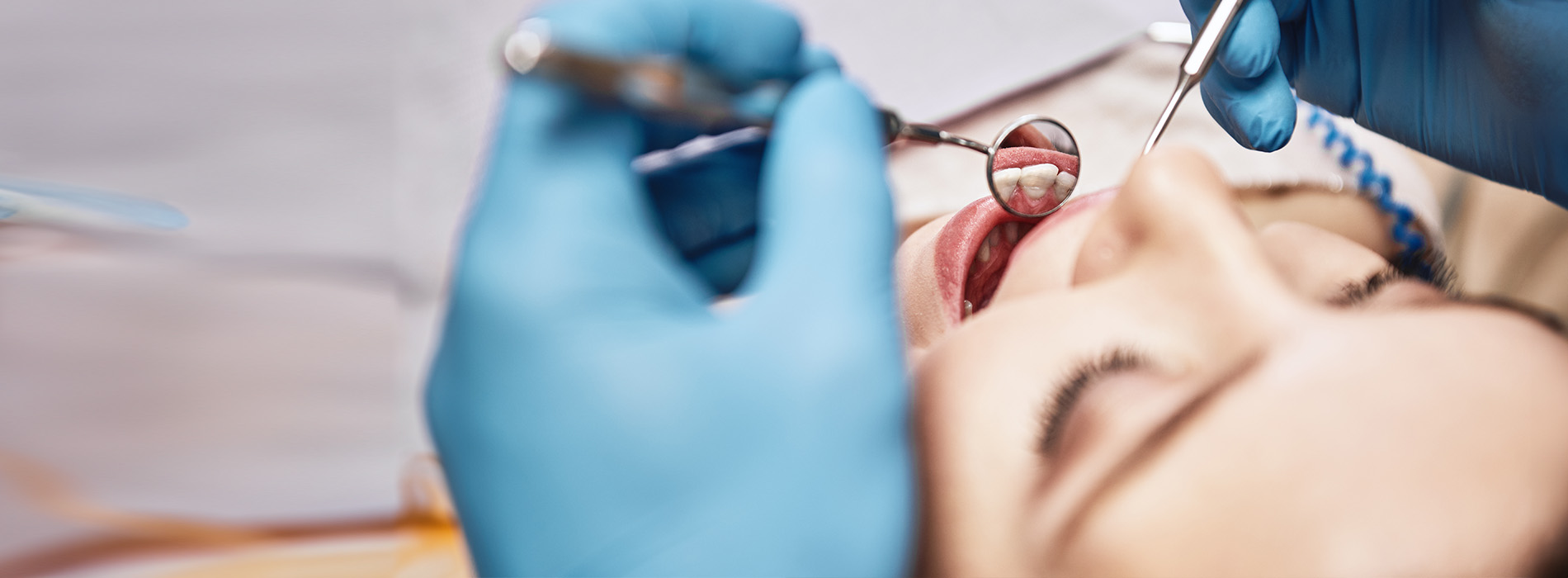 The image shows a person receiving dental care, with a dentist performing a procedure, using tools such as a drill and a mirror, while wearing protective gloves and an apron.