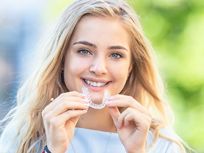 The image features a young woman smiling at the camera while holding a clear retainer in her hand.