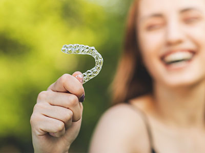 The image shows a young woman holding up an object shaped like a smiley face with her right hand, while she appears to be laughing or smiling broadly at the camera.