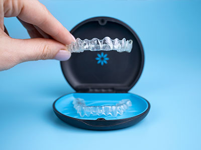 The image shows a person s hand holding an open case containing a set of clear aligners, which are used for teeth straightening, with the aligners partially inserted into a blue mouthguard-like device.