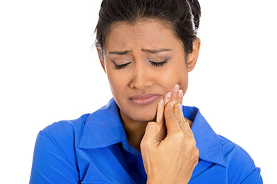 The image shows a woman with her eyes closed, looking upwards, seemingly in distress or pain, holding her hand near her face where she appears to be experiencing discomfort or irritation, possibly related to dental health, against a plain background.