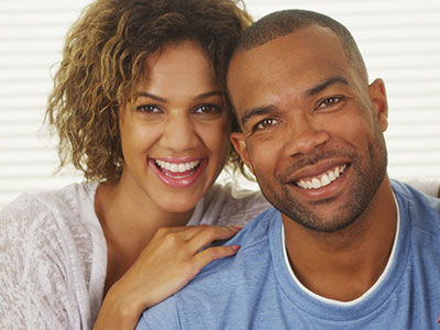 A man and woman are smiling at the camera  the man has short hair and is wearing a blue t-shirt, while the woman has long hair and is wearing a white top.