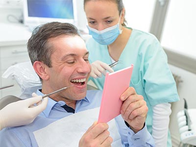 A smiling man holding a pink card with a surprised expression while sitting in a dental chair surrounded by dental equipment and a female dentist.