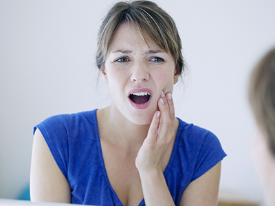 A distressed woman with her mouth open, looking at her reflection in a mirror, with her hand on her chin.