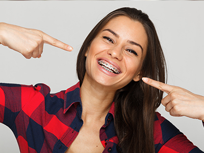 A woman with a wide smile pointing at her teeth, wearing a red plaid shirt and posing playfully for the camera.