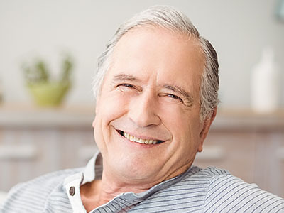 The image shows an elderly man with gray hair smiling at the camera while sitting comfortably indoors.